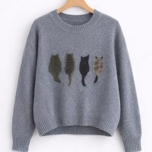 Könnte beinhalten: Ein grauer Strickpullover mit Rundhalsausschnitt und gerippten Bündchen und Saum. Der Pullover hat vier gestickte Katzensilhouetten in Grün-, Braun- und Schwarztönen auf der Brust. Der Pullover hängt an einem Holzkleiderbügel.