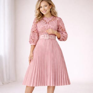 May include: A pink dress with a pleated skirt and a matching belt. The top of the dress has a lace design with three-quarter sleeves. The belt has a silver buckle. The dress is a light shade of pink.