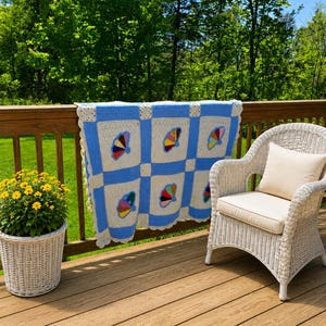 May include: A handmade crochet blanket featuring a grid pattern of light blue and cream-colored squares. Each cream square contains a colorful fan-shaped motif in shades of red, yellow, blue, and black. The blanket has a scalloped white border.