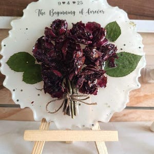 Könnte beinhalten: Ein weißer Resin-Untersetzer mit einem Strauß getrockneter burgunderfarbener Rosen und grünen Blättern. Der Untersetzer trägt den Text "9 4 25" und "The Beginning of forever". Er wird auf einer kleinen Holzstaffelei präsentiert.
