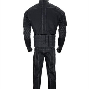 May include: A black tactical-style jumpsuit featuring a high collar, long sleeves with white geometric grid stitching on the forearms, a padded waist belt, and white contrast stitching throughout the garment, displayed on a mannequin.