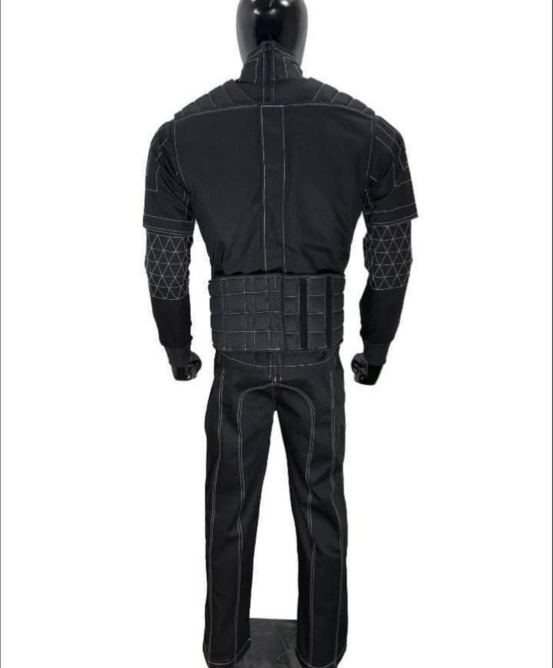 May include: A black tactical-style jumpsuit featuring a high collar, long sleeves with white geometric grid stitching on the forearms, a padded waist belt, and white contrast stitching throughout the garment, displayed on a mannequin.
