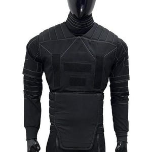 May include: A black tactical-style long-sleeve shirt and vest set featuring a high mock neck collar, padded shoulder panels, and a structured midsection. The garment is accented with white contrast stitching throughout and rectangular velcro patches.
