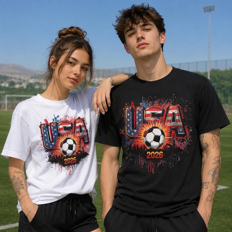 Pu&ograve; includere: Due persone indossano magliette abbinate con un design a tema calcistico. Le magliette presentano la scritta "USA" in rosso, bianco e blu, con un pallone da calcio e l'anno "2026" stampato sotto. Una maglietta &egrave; bianca, l'altra &egrave; nera.