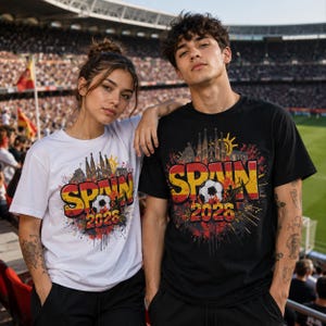 Pu&ograve; includere: Due persone indossano magliette con un design Spagna 2026. Le magliette presentano la parola "SPAIN" in giallo e rosso, un pallone da calcio e una grafica di uno skyline. Una maglietta &egrave; bianca, l'altra &egrave; nera.
