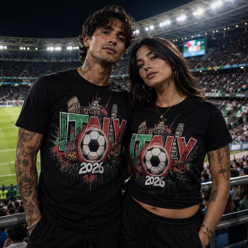 Pu&ograve; includere: Magliette nere con grafica di pallone da calcio e la scritta "ITALY" in verde e rosso, con l'anno "2026". Le magliette presentano monumenti italiani. L'immagine mostra due persone che indossano le magliette.