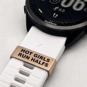 May include: A black Garmin smartwatch with a white band. The watch face displays the time, battery life, and running data. A gold-colored band keeper with the text "HOT GIRLS RUN HALFS" is on the white band.