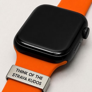 May include: Black smartwatch with an orange band. A silver band keeper displays the text "THINK OF THE STRAVA KUDOS". The watch has a black screen and a black side button. The band is made of a flexible material.