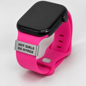 May include: A black smartwatch with a bright pink band. The band features a silver metal plate with the text "HOT GIRLS DO HYROX". The watch has a black screen and a black dial on the side.