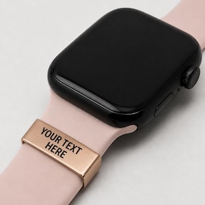 May include: A black smartwatch with a pale pink strap. A gold-coloured metal band with the text "YOUR TEXT HERE" is attached to the watch strap. The watch is displayed on a light grey surface.