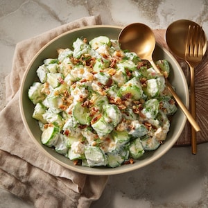 Peut inclure: Une salade de concombres fraîche dans un bol vert, composée de morceaux de concombres, d'une sauce crémeuse et de morceaux de bacon croustillants. Garnie d'aneth frais, la salade est servie avec des ustensiles dorés.