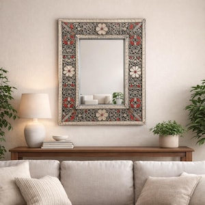 May include: Rectangular mirror with a decorative frame featuring floral and geometric patterns in red, white, and black. The mirror is mounted on a light-colored wall above a wooden console table. A lamp and plants flank the mirror.