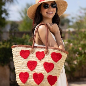 May include: A woven straw tote bag with red heart appliqués. The bag has brown leather handles and trim. The person in the image is wearing a straw hat with a red band and sunglasses.