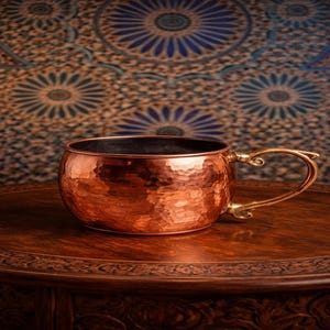 Peut inclure: Un mug Moscow Mule en cuivre avec une finition martelée et une poignée en laiton. Le mug est posé sur une table en bois foncé avec un bord décoratif, sur un fond à motifs.