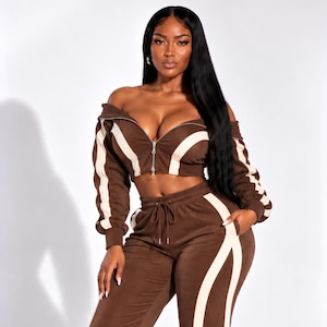 May include: A brown two-piece set consisting of a cropped, off-the-shoulder zip-up jacket and wide-leg pants. Both pieces feature cream-colored vertical stripes along the sleeves and legs. The jacket has a silver zipper and the pants have a drawstring.