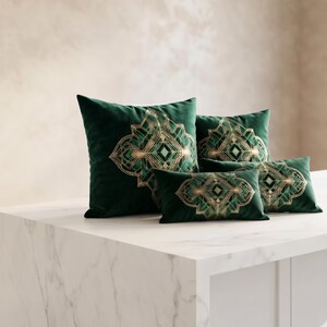 May include: A collection of four dark green velvet throw pillows in varying sizes, each featuring a symmetrical gold and light green geometric pattern centered on the front. The pillows are arranged in a cluster, showcasing square and rectangular shapes.