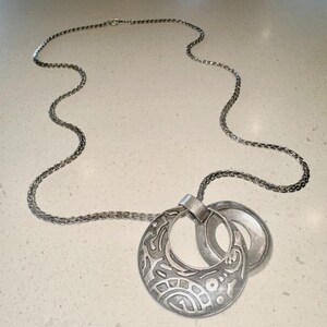 May include: Silver-toned necklace featuring a long chain and a pendant with two connected circular elements. The larger element has an open center and an etched, patterned design. The smaller element is a plain ring.