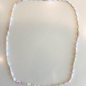 May include: A pastel-colored beaded necklace with a silver clasp. The necklace features rectangular beads in shades of pink, purple, yellow, and green. The beads are strung together to form a long, elegant chain.