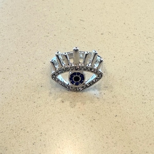 Puede incluir: Anillo plateado con diseño de ojo. El anillo tiene un centro azul y está rodeado de cristales transparentes. El ojo está enmarcado por una banda plateada con detalles verticales, creando un accesorio único y elegante.
