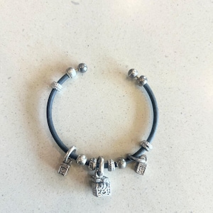 May include: A dark gray bracelet with silver charms. The bracelet has a dark gray band with silver beads and three square charms. The charms have intricate designs. The bracelet is on a light gray surface.