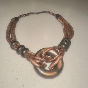 May include: A copper-colored necklace with a large, circular pendant. The necklace features multiple strands of small, round beads and dark brown accents. The pendant is a large, smooth ring. The necklace is designed to be worn around the neck.
