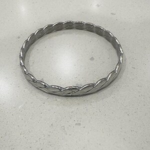 May include: A silver-colored bangle bracelet with a braided design. The bracelet is circular and appears to be made of metal. The braided pattern gives the bracelet a textured appearance. The bracelet is displayed on a light-colored surface.
