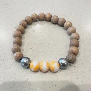 May include: A beaded bracelet featuring light brown wooden beads and a series of gray and yellow-white beads. The bracelet is made of round beads strung together, creating a circular shape. The bracelet is on a light gray surface.