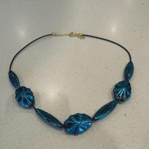 May include: A necklace featuring iridescent blue beads in various shapes, including flower-like and oval designs. The beads are strung on a black cord with a gold-colored clasp. The necklace is a jewelry item.