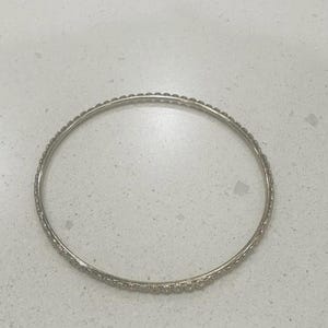 May include: A silver-colored bangle bracelet with a textured surface. The bracelet is circular and appears to be made of metal. The design features small, evenly spaced details around the band, adding a touch of elegance to the jewelry.