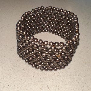 May include: A bronze-colored beaded bracelet. The bracelet is made of many small, round beads strung together to form a wide, cuff-style design. The beads have a metallic sheen, reflecting light. This jewelry piece is suitable for everyday wear or special occasions.