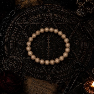 May include: A circular bracelet composed of numerous small, textured, gold-toned spherical beads. The bracelet is centered on a dark, circular surface featuring intricate engraved geometric patterns, occult-style symbols, and printed text.