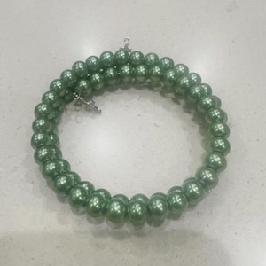 Pode incluir: Uma pulseira em espiral composta por contas verdes e redondas. As contas são enfiadas, formando um design flexível e enrolado. A pulseira tem um fecho prateado. As contas têm um acabamento brilhante, que reflete a luz.