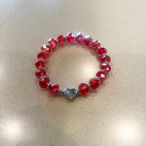 May include: A red beaded bracelet with a silver heart charm. The bracelet features faceted red beads and silver accents. The heart charm is a solid silver colour. The bracelet is circular and sits on a light-coloured surface.