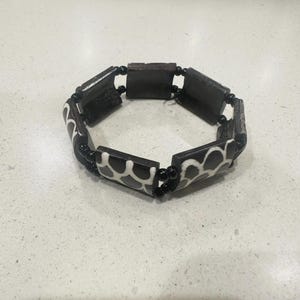 May include: A black and white bracelet with a geometric pattern. The bracelet is made of rectangular wooden beads and black spherical beads. The design features a repeating pattern of white shapes on a black background.