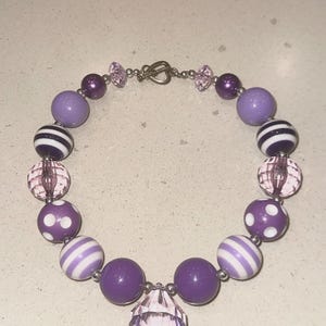 May include: A purple beaded necklace with various bead styles. The necklace features large, round purple beads, striped beads, and polka dot beads. A large, faceted crystal pendant hangs from the centre. The necklace is approximately 40 cm in length.