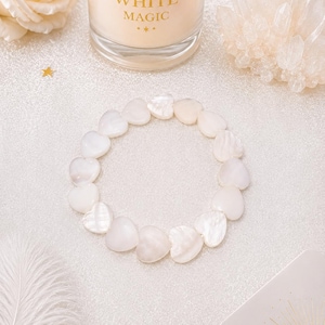 Puede incluir: Una pulsera circular compuesta por cuentas blancas en forma de corazón, lisas e iridiscentes. Las cuentas presentan un brillo nacarado y están ensartadas para formar un bucle continuo, mostrando un acabado pulido que refleja la luz.
