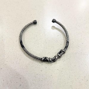 May include: A silver-toned cuff bracelet with a twisted rope design. The bracelet features a series of intertwined loops and small embellishments. The ends of the bracelet have decorative caps. Jewelry.