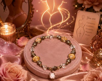 May include: A beaded necklace featuring alternating clear and amber-colored faceted crystals, metallic textured spacers, and a central oval pendant. The necklace is displayed on a circular velvet cushion surrounded by pink roses and candles.