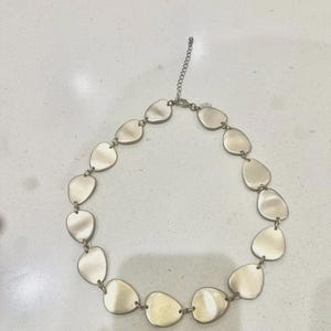 May include: A silver necklace with a series of connected, irregularly shaped, flat pendants. The pendants are smooth and have a slightly reflective surface. The necklace has a chain extender for adjustable length.