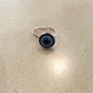 May include: A silver-coloured adjustable ring with a circular charm displaying a blue and white evil eye design. The ring is set against a neutral background, emphasising the ring's details.