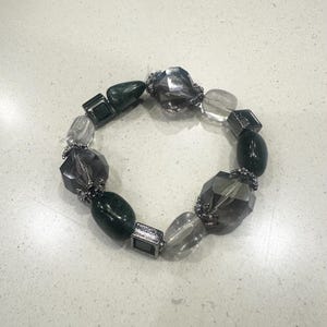 May include: A beaded bracelet featuring a mix of dark green, clear, and smoky gray faceted beads. The bracelet includes square and oval-shaped beads, with silver-toned metal accents. The beads are strung together to form a circular shape.