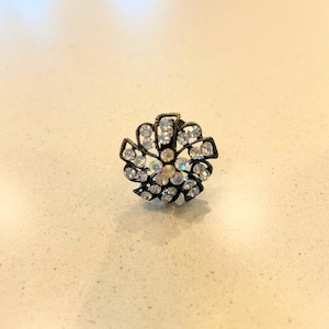 May include: A floral-shaped ring with a dark metal base and clear, sparkling crystals. The ring's design features a central cluster of crystals surrounded by petals of smaller stones. The crystals catch the light, creating a dazzling effect.