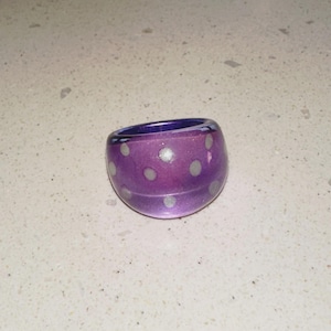 Peut inclure: Une bague en verre violet épais de forme arrondie. La bague est ornée de petits pois blancs espacés. La bague est posée sur une surface de couleur claire.