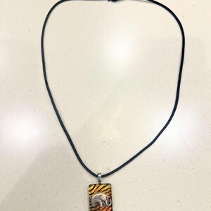 May include: A black cord necklace with a rectangular pendant featuring a silver elephant design. The pendant has a colourful background with orange, yellow, and black stripes. The necklace is displayed on a light-coloured surface.