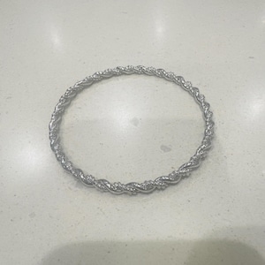 Könnte beinhalten: Ein silberfarbenes Armreif-Armband mit einem gedrehten Seildesign. Das kreisförmige Armband besteht aus Metall und weist kleine dekorative Elemente entlang des gedrehten Musters auf. Das Armband wird auf einer hellen Oberfläche präsentiert.