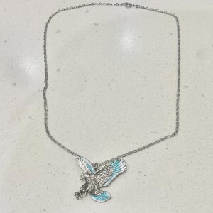Puede incluir: Collar plateado con un colgante de águila con las alas extendidas. El águila tiene detalles de plumas y acentos de esmalte azul en las alas. La cadena es delicada y el colgante está centrado.