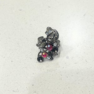 May include: A dark metal ring with an ornate design featuring a cluster of colourful rhinestones. The ring has a floral motif with clear, pink, and dark grey stones. The ring is set against a white background.