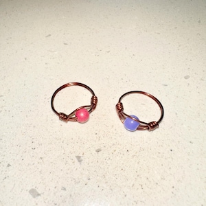 May include: Two handmade wire rings. One ring features a pink bead, and the other has a blue bead. The rings are crafted with copper-colored wire, with the beads centered in the design. The rings are displayed on a white surface.
