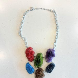 Könnte beinhalten: Eine silberne Halskette mit sieben bunten, unregelmäßig geformten Edelsteinanhängern. Die Anhänger sind in den Farbtönen Rot, Grün, Lila, Blau, Braun und Schwarz gehalten. Die Halskette wird auf einer weißen Oberfläche präsentiert.