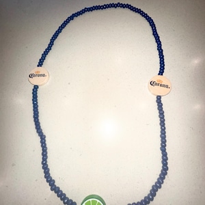 Puede incluir: Un collar de cuentas azules con un colgante de rodaja de lima y dos colgantes de madera con el logotipo de Corona. La rodaja de lima es verde con detalles blancos. Los colgantes de Corona son circulares.